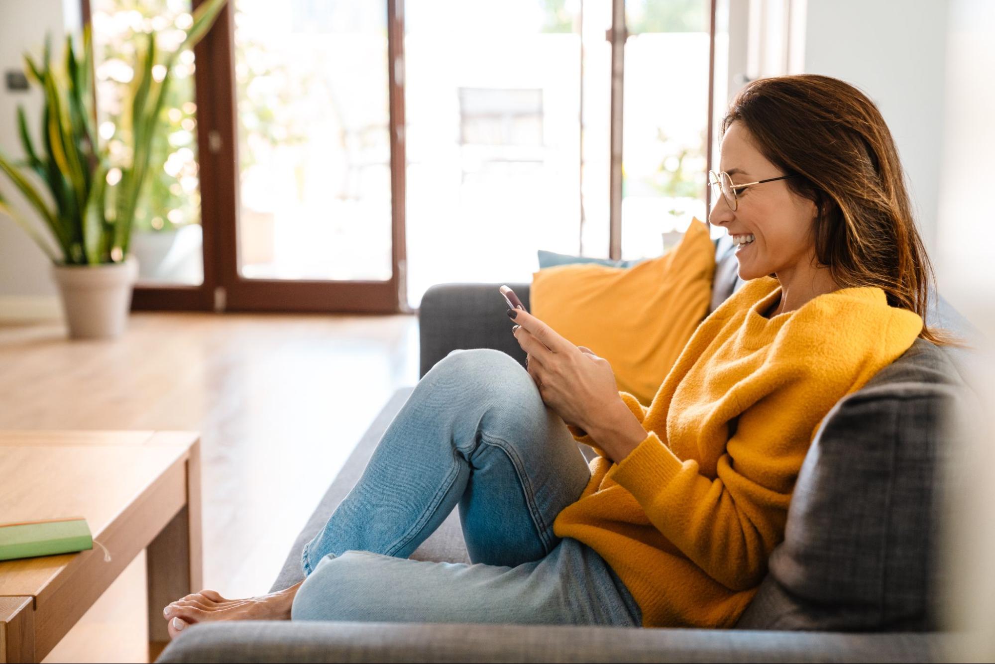 Mulher de blusa amarela e calça jeans mexe em celular sentada no sofá de casa. Imagem ilustrativa para post sobre nova ferramenta de gestão de imóveis desenvolvida pela Soma Urbanismo.